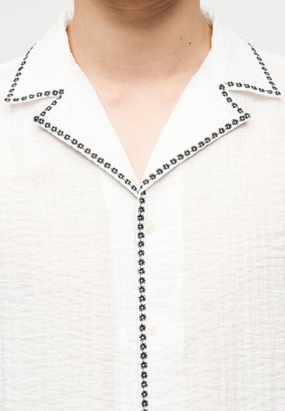 Λευκό πουκάμισο με υφή και μαύρη κεντητή διακόσμηση λουλουδιών που περιγράφει το γιακά και την ταινία κουμπιών στο πάνω μέρος του στήθους και του λαιμού ενός ατόμου.