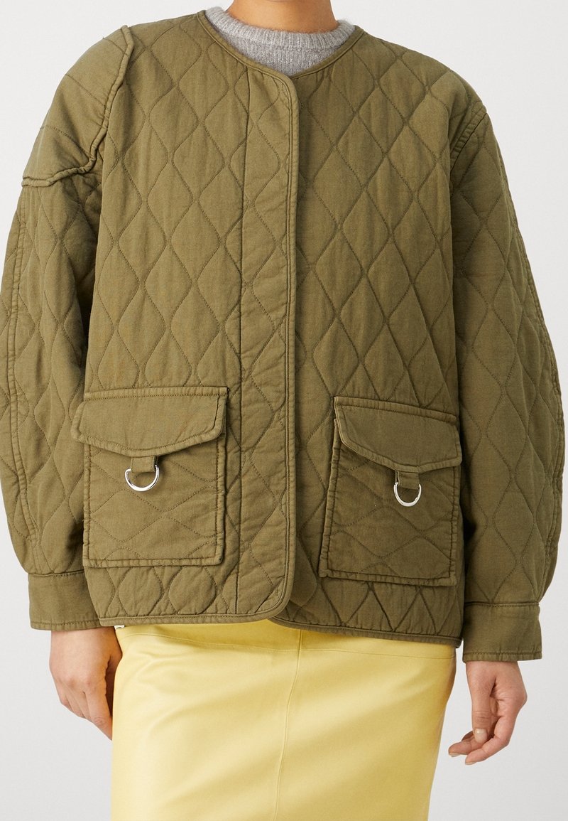 Personne portant une veste matelassée vert olive avec poches à rabat et anneaux argentés, associée à une jupe jaune clair.