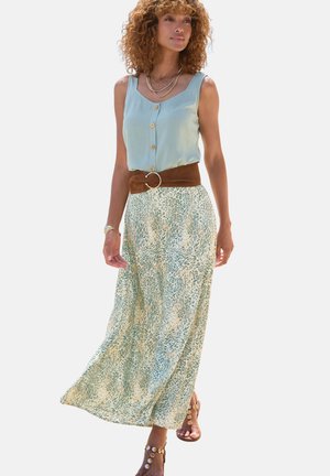 Frau trägt ein ärmelloses hellblaues Oberteil mit Knöpfen, einen gemusterten langen Rock, einen breiten braunen Gürtel und Sandalen mit Knöchelriemen und Goldmünzen.