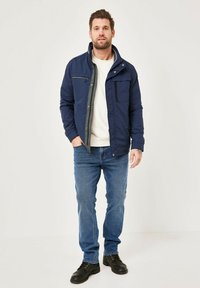 Navy-blue insulated jacket with a high collar, front zip, and multiple pockets. Worn over a light sweater and paired with blue jeans and black shoes.
