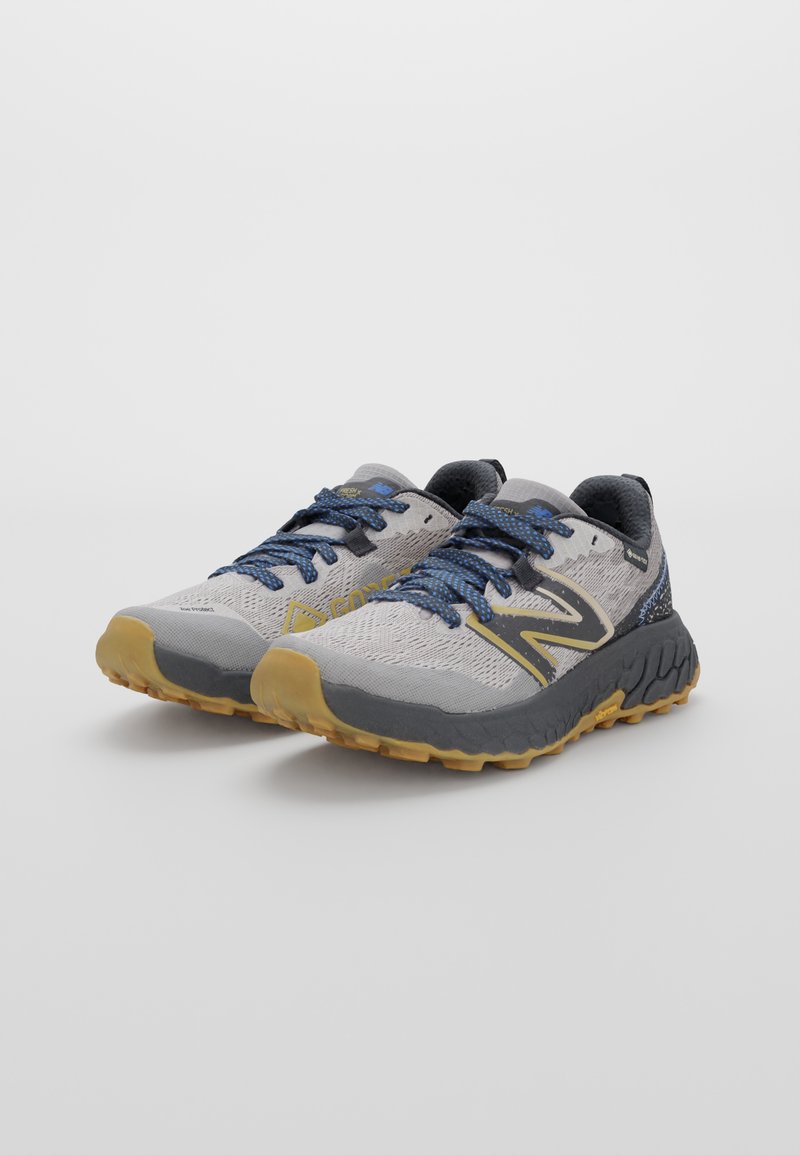 Zapatillas de trail running grises y amarillas con parte superior de malla texturizada, cordones azules y suela de goma con un patrón de tracción profundo para agarre.