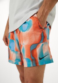 Homme portant un short à motif abstrait orange, bleu et blanc avec logo Nike noir, main dans la poche et haut gris clair texturé.