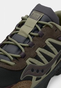 Běžecká obuv Saucony s šedou a hnědou horní částí, texturovaným materiálem, zelenými tkaničkami a detailem loga, opatřená polstrovaným límcem.