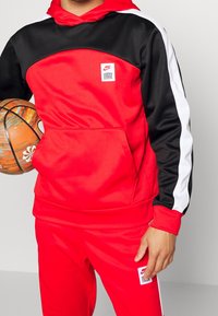 Sweat à capuche rouge et noir avec une poche avant, des bandes blanches sur les côtés, et un petit logo Nike. Porté avec un pantalon rouge assorti, tenant un ballon de basket.