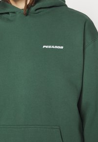 Grön huvtröja i mjukt material med en framficka och vitt broderat "PEGADOR"-logotyp på bröstet.