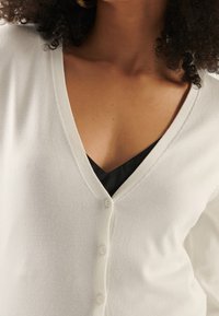 Vit k cardigan i mjukt tyg med djup V-ringning, förestående av fem knappar fram och en slät yta.