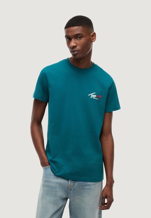 Młody mężczyzna ubrany w morski t-shirt Tommy Jeans oraz jasnoniebieskie dżinsy, z jedną ręką w kieszeni, stoi na jednolitym tle.