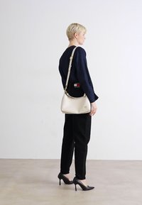 Beige leather handbag with a smooth texture, curved shape, gold-tone hardware, and an adjustable strap. Worn crossbody against a black outfit.