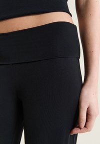 Gros plan sur une personne portant un legging noir taille haute et un haut court noir, la main droite reposant à côté de la jambe.