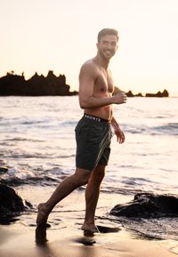 Groene zwembroek met een elastische tailleband en trekkoord. Het model is blootsvoets en loopt langs een strand met golven en rotsen.