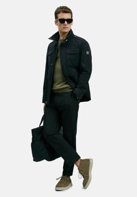Man in zwarte jas, olijfgroene trui, zwarte broek, tan suède schoenen, zonnebril, met een zwarte tas tegen een witte achtergrond.