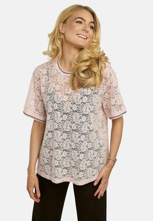 Femme souriante aux longs cheveux blonds portant un haut en dentelle rose clair à manches courtes et un pantalon noir, debout devant un fond blanc.