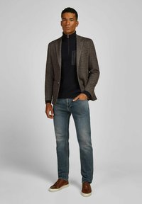 Man standing wearing brown textured blazer, black quarter-zip sweater, blue jeans, and brown sneakers against plain light background.