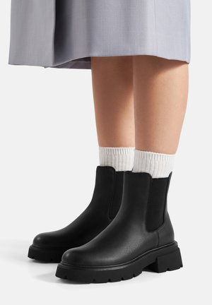 Stivaletti Chelsea neri in pelle liscia con pannelli elastici laterali a contrasto e suole di gomma spesse. Calzini bianchi a coste visibili sopra gli stivaletti.