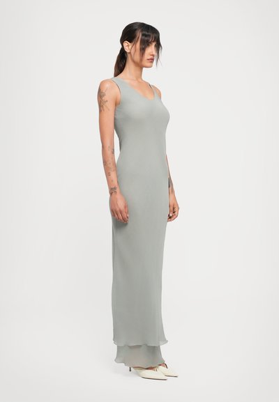 Woman standing sideways in a sleeveless, light gray, floor-length dress with white pointed heels and visible arm tattoos.
