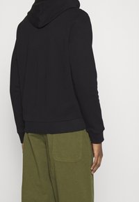 Sweat à capuche en coton noir avec une capuche spacieuse et des poignets côtelés, associé à un pantalon ample vert olive avec des poches sur le côté et à l'arrière.