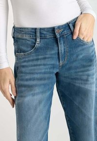 Jean en denim bleu avec une coupe classique, doté d'une fermeture par bouton à l'avant, de cinq poches et de détails de frottement subtils. Texture lisse.