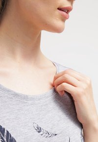 Šedé tričko s krátkým rukávem a kulatým výstřihem, potisk s peřím; ženská ruka jemně drží límec trička blízko výstřihu.