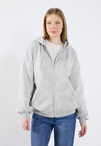 Felpa leggera grigia con zip, realizzata in tessuto morbido. Presenta grandi tasche anteriori, cappuccio con cordino e polsini e orlo a costine. Abbinata a jeans blu.