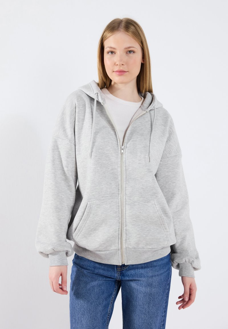 Felpa leggera grigia con zip, realizzata in tessuto morbido. Presenta grandi tasche anteriori, cappuccio con cordino e polsini e orlo a costine. Abbinata a jeans blu.
