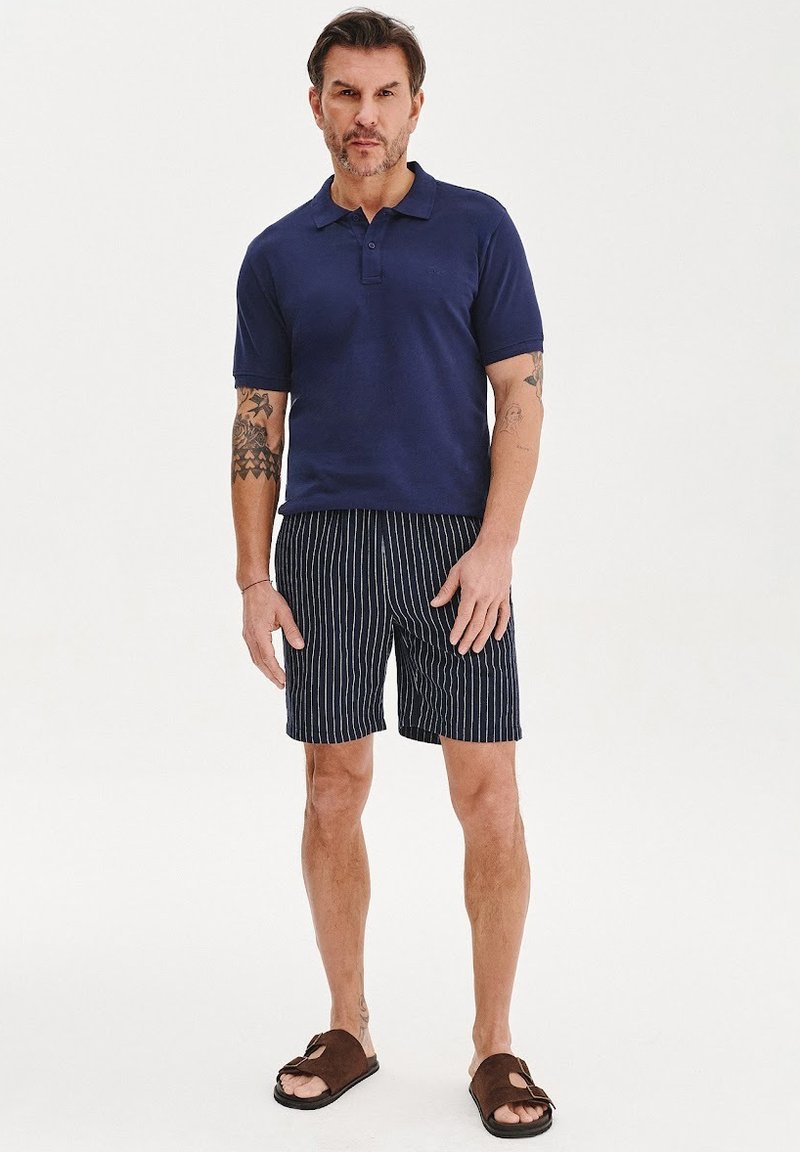 Granatowa koszulka polo, w paski granatowe szorty i brązowe sandały. Strój charakteryzuje się klasycznym wzornictwem, miękką teksturą i swobodnym krojem.