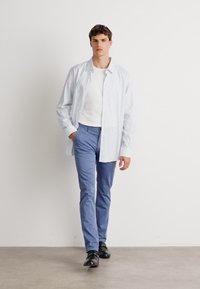 Chemise boutonnée rayée bleu clair, t-shirt blanc, pantalon bleu ajusté et chaussures noires. Le mannequin se tient devant un fond blanc.