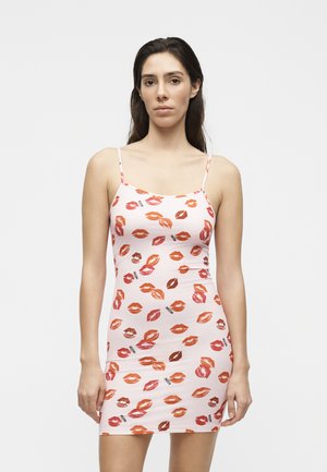 Nő, aki egy ujjatlan rózsaszín miniruhát visel, piros és narancssárga ajaknyomatos mintákkal, fehér háttér előtt áll.