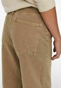 Pantalons taille haute de couleur brun clair, en tissu texturé, avec deux poches arrière et des coutures visibles le long des côtés.