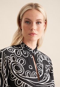 Schwarze Bluse mit weißen Blumendrucken, hoher Kragen mit Rüschendetail, Knopfverschluss und einem Ausschnitt mit Schlüsselelement. Weicher, strukturierter Stoff.