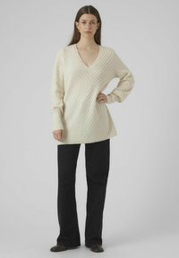 Beige knitted sweater with a V-neck and chevron pattern, featuring long sleeves and a relaxed fit. Paired with black flared pants.