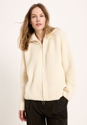 Woman with light brown hair wearing a cream ribbed zip-up sweater and black pants, standing against a plain light background.