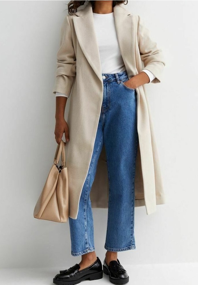 Abrigo largo beige con solapas, camisa blanca, jeans azules de cintura alta y mocasines negros; sostiene un bolso texturizado beige.