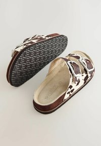 Bruna och vita ko-prickiga slidesandaler med två metallspännen, ovandel av mocka och texturerad svart gummisula. Lätta och öppna i designen.