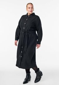 Naine kannab pikka musta nööbiga kleiti, millel on vöökohaga pael, ja musti ankle-boote kaunistavate rihmadega, seistes lihtsa tausta ees.