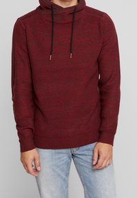 Roter, gestrickter Pullover mit einem Schalkragen, schwarzen Kordelzügen, gerippten Bündchen und einer lockeren Passform. Getragen über hellblauen Jeans.