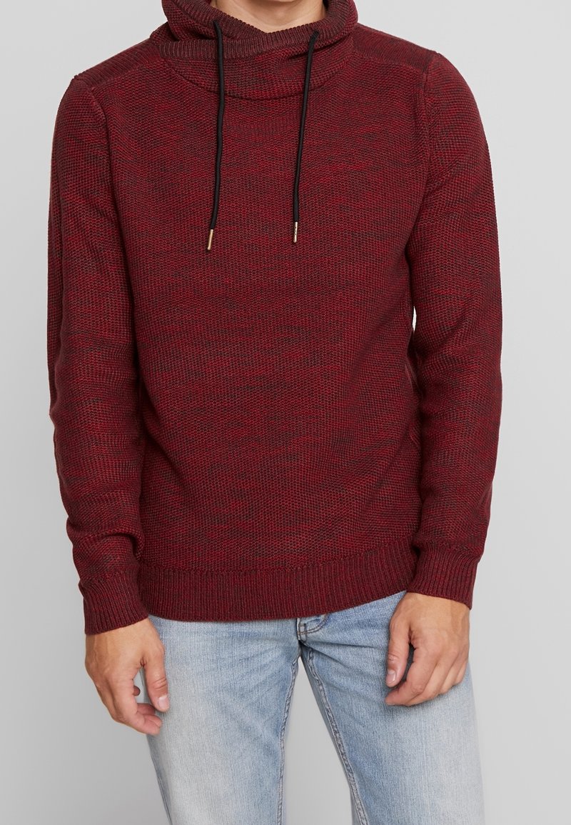 Roter, gestrickter Pullover mit einem Schalkragen, schwarzen Kordelzügen, gerippten Bündchen und einer lockeren Passform. Getragen über hellblauen Jeans.