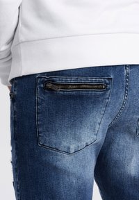 Tummansinisiä farkkuja, joissa on vetoketjullinen takatasku, haalistunut pinta ja muotilöydöt, yhdistettynä valkoiseen ribatutuuriin.