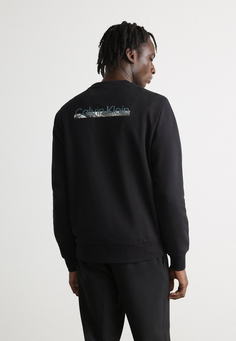 Zwarte sweatshirt met een ronde hals en ribgebreide manchetten. De achterkant toont het "Calvin Klein" logo in het blauw met een getextureerde grafische accenten.