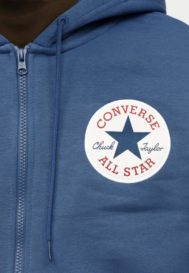 Blå hoodie med lynlås med et cirkulært hvidt mærke med "CONVERSE" og "ALL STAR" i rødt, en marineblå stjerne og teksten "Chuck Taylor." Blød tekstur.