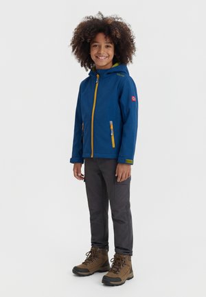 Smiling child with curly hair wearing a blue hooded jacket with yellow zippers, dark pants, and brown hiking boots, standing relaxed.