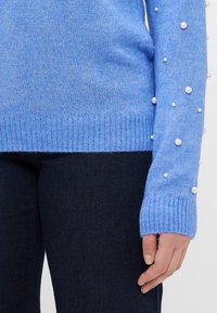 Lyseblå strikkegenser med perledetaljer langs ermekantene, ribbestrikket fald og teksturert finish. Sammen med mørke jeans.