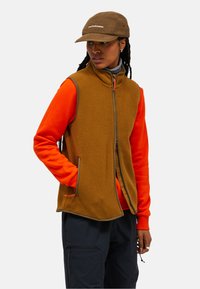 Brauner Fleece-Weste mit hohem Kragen und Reißverschluss, getragen über einem orangefarbenen Langarmshirt. Akzentuiert mit dunklen Besätzen und einem strukturierten Stoff.