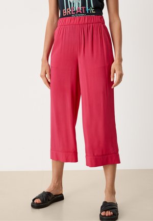 Žena oblečená v volných, zkrácených červených kalhotách s elastickým pasem a černých nazouvacích sandálech, stojící proti jednobarevnému pozadí.