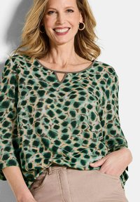 Blusa verde con stampa leopardata, dal tessuto morbido, con scollatura rotonda e un piccolo dettaglio a chiave davanti, abbinata a pantaloni beige.