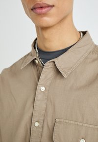 Beige skjorte med knaplukning og struktureret stof, en enkelt brystlomme og standard krave, lagdelt over en mørkegrå top. Synlig sølvkæde.