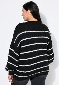 Frau mit langen dunklen Haaren, die große Creolen und einen schwarzen Strickpullover mit weißen horizontalen Streifen trägt, steht an einer weißen Wand.
