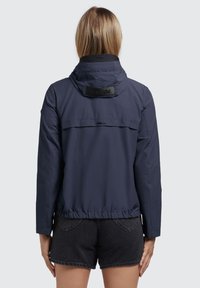 Marine windbreaker jack met capuchon, gathered zoom en details in de achterzak. Kenmerken zijn het merklogo op de nek en elastische manchetten.