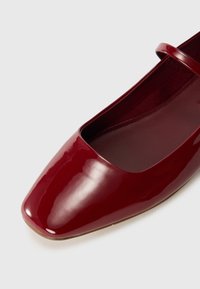 Chaussure plate en cuir verni rouge avec un bout carré, une lanière simple et une finition lisse. La doublure intérieure est rouge foncé, offrant un contraste.