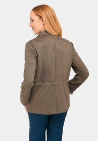 Braune Wildlederjacke mit taillierter Passform, Gürtel in der Taille, langen Ärmeln und Rückennaht-Details. Getragen über blauen Jeans.