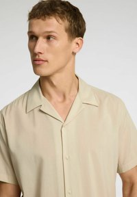 Camicia beige a maniche corte realizzata in tessuto leggero. Presenta un colletto classico, una chiusura con bottoni e una texture liscia.